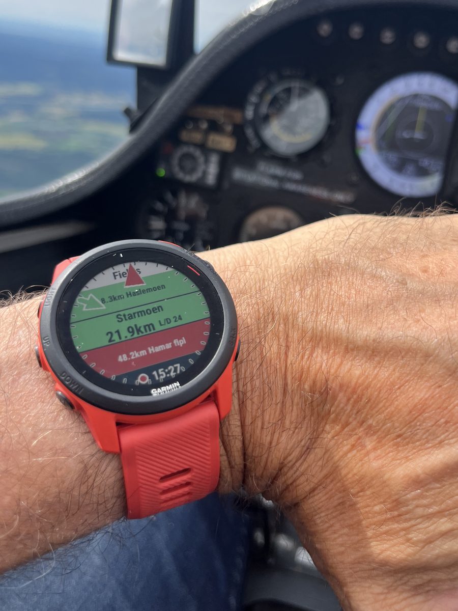 A red Garmin Forerunner on the wrist showing GlideRange — Starmoen 21.9 km within glide — beside a glider's instrument panel in flight over Norwegian terrain.