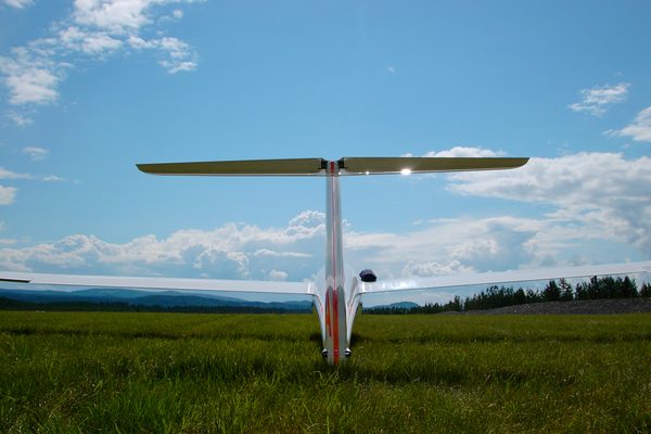 A glider seen head-on from behind on a green summer meadow, wings level and cumulus building over the hills.